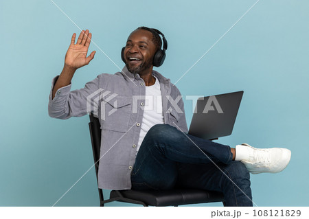Excited black man with headphones using laptop looking away waving hand saying hello Excited black man with headphones using laptop looking away waving hand saying hello 108121829