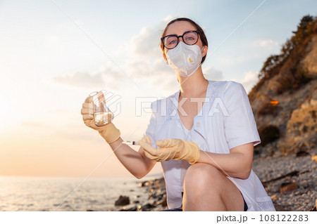 Portrait of young woman scientist-ecologist wearing eyeglasses, mask and rubber gloves examines flask of water from ocean. Copy space. Concept of ecology and science Portrait of young woman scientist-ecologist wearing eyeglasses, mask and rubber gloves examines flask of water from ocean. Copy space. Concept of ecology and science 108122283