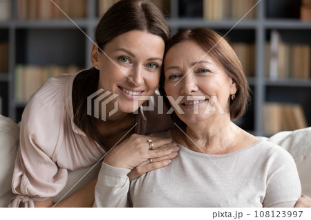 Adult daughter hugs from behind seated on sofa elderly mother Adult daughter hugs from behind seated on sofa elderly mother 108123997