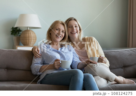 Happy blonde senior mom and adult daughter drinking hot beverage Happy blonde senior mom and adult daughter drinking hot beverage 108124275