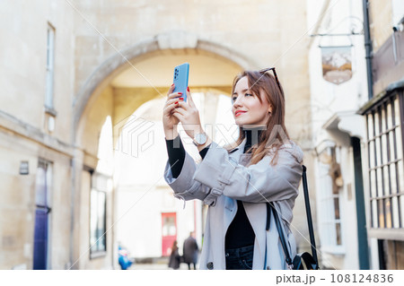 Young tourist woman visiting a picturesque city street destination, using smart phone taking pictures during holiday and travel walk in old European city, outdoors. Sightseeing trip through the city. 108124836