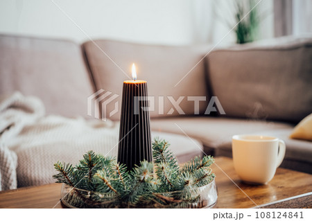 Composition of flaming black christmas candle and spruce branches and cup with hot drink on coffee table in living room. Natural Christmas decorations for home. Cozy Christmas winter domestic mood. Composition of flaming black christmas candle and spruce branches and cup with hot drink on coffee table in living room. Natural Christmas decorations for home. Cozy Christmas winter domestic mood. 108124841