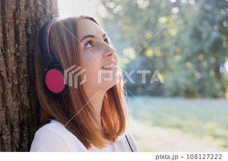Portrait of red haired teenage girl listening to music in big pink earphones outdoors leaning to a tree. Portrait of red haired teenage girl listening to music in big pink earphones outdoors leaning to a tree. 108127222