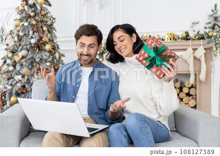 Smiling young couple man and woman talking from home on video call from laptop. They are sitting on the sofa, holding a gift in their hands. Smiling young couple man and woman talking from home on video call from laptop. They are sitting on the sofa, holding a gift in their hands. 108127389