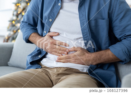Close-up photo of young man's hands holding his stomach. A man sits on a couch and suffers from pain, spasms, poisoning, overeating. Close-up photo of young man's hands holding his stomach. A man sits on a couch and suffers from pain, spasms, poisoning, overeating. 108127396
