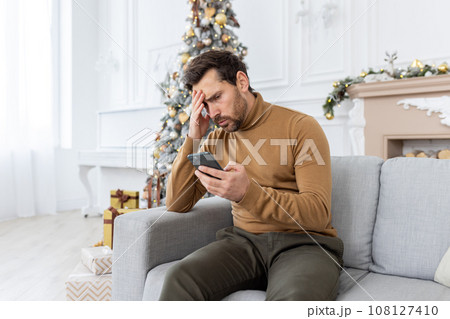 A young man sitting at home on the sofa received bad news on the phone on the eve of the New Year and Christmas holidays. Disappointed and shocked, he leaned his head on his hand. 108127410