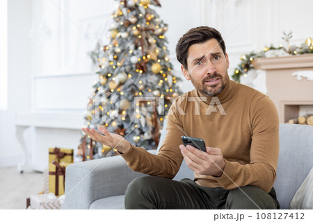 Portrait of a young man sitting at home on New Year's holidays near the Christmas tree on the sofa, using the phone and unhappily waving his hands at the camera. 108127412