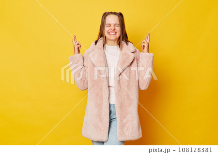Smiling young girl with funny ponytails standing isolated over yellow background holding fingers crossed for good luck having desire closed eyes. Smiling young girl with funny ponytails standing isolated over yellow background holding fingers crossed for good luck having desire closed eyes. 108128381
