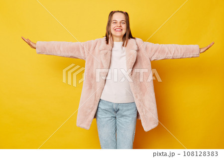 Charming adorable woman dressed pink fur coat isolated over yellow background spreading arms pretending flying looking smiling at camera expressing happiness. Charming adorable woman dressed pink fur coat isolated over yellow background spreading arms pretending flying looking smiling at camera expressing happiness. 108128383