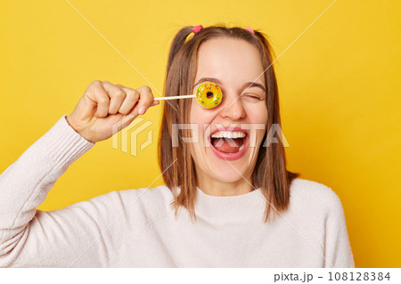 Caucasian adult smiling woman covering her eye with lollipop isolated over yellow background smiling to camera having excited amazed facial expression. Caucasian adult smiling woman covering her eye with lollipop isolated over yellow background smiling to camera having excited amazed facial expression. 108128384