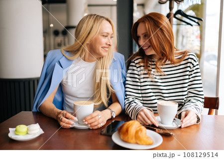 Portrait of cheerful young best girlfriends sitting at modern cafe, communicating and sharing stories, spending relax in coffee shop. Two women friends sitting in cafeteria and talking. 108129514