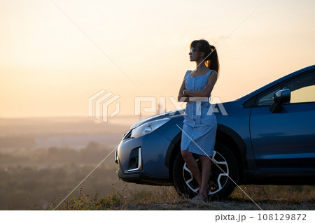 Happy woman driver in blue summer dress enjoying warm evening near her car. Travel and vacations concept. Happy woman driver in blue summer dress enjoying warm evening near her car. Travel and vacations concept. 108129872