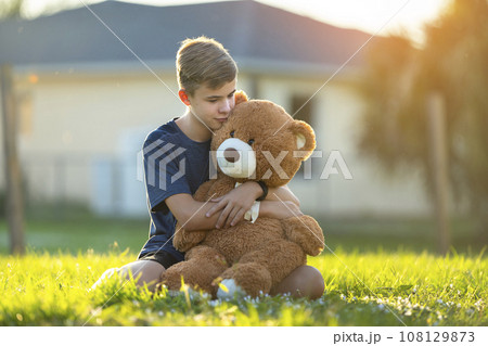Happy teenager boy hugging his teddy bear friend outdoors on green grass lawn. Friendship concept 108129873