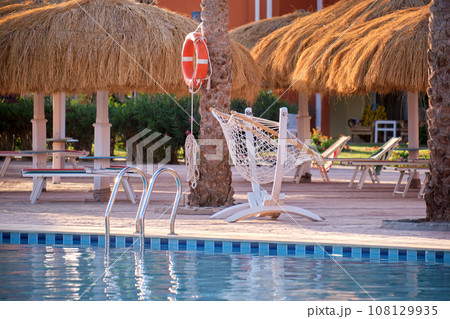 Empty deck chairs under straw shade umbrellas on swimming pool side in tropical resort. Summer vacations and getaway concept Empty deck chairs under straw shade umbrellas on swimming pool side in tropical resort. Summer vacations and getaway concept 108129935