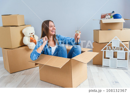 Happy woman drinks champagne from a glass while sitting inside a cardboard box in new apartment. Happy woman drinks champagne from a glass while sitting inside a cardboard box in new apartment. 108132367