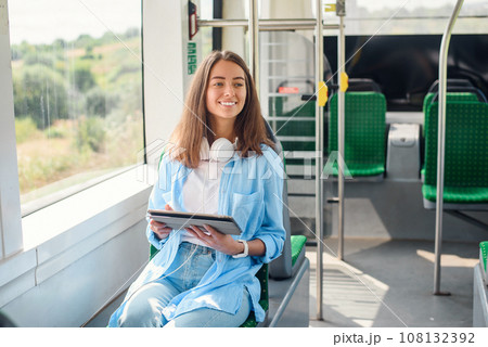 Happy smiling woman reads a tablet or ebook in a modern tram or subway. Pretty girl rides for work in public transport. 108132392