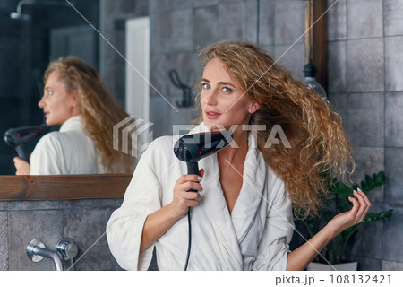 Close up of appealing smiling young lady in white dressing gown posing on camera while drying her curly hair using hair dryer near the bathroom mirror 108132421