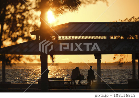 Dark silhouette of people resting under alcove roof on sea shore in public park at sunset Dark silhouette of people resting under alcove roof on sea shore in public park at sunset 108136815