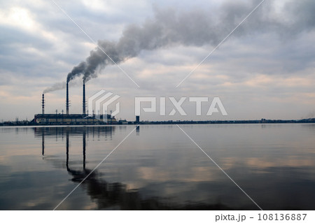 Coal power plant high pipes with black smoke moving upwards polluting atmosphere with reflections of it in lake water. Coal power plant high pipes with black smoke moving upwards polluting atmosphere with reflections of it in lake water. 108136887