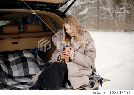 Young blonde girl sitting in an open car's trunk. Girl holding thermos with tea in hands. Teenager wearing beige jacket. Young blonde girl sitting in an open car's trunk. Girl holding thermos with tea in hands. Teenager wearing beige jacket. 108137451