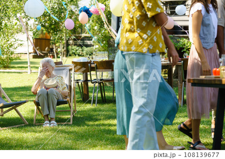 Tired elderly woman sitting on folding chair at garden party, taking break after long outdoor grill party. Elderly woman cries tears of joy, happy to be with her family and loved ones. 108139677