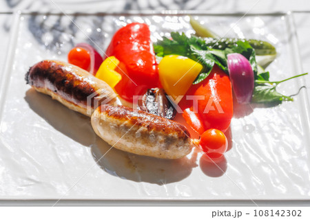 grilled sausages with vegetables on a white plate on a sunny summer day 108142302