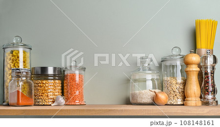 Various uncooked groceries and pasta in glass jars arranged on wooden shelf on olive green wall. 108148161