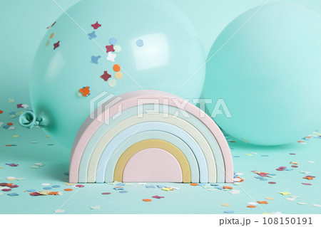a plastic rainbow toy in front of balloons of the same color as the background and confetti on the ground. 108150191