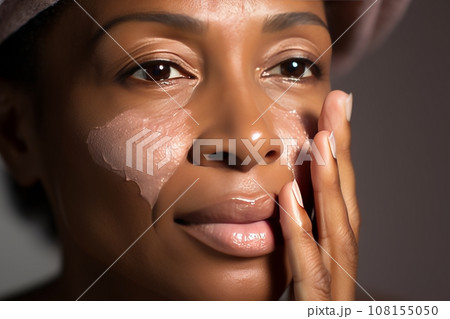 Close-up of middle-aged African American woman touching her face to apply moisturizer. Smiling face of adult colored lady with daily cream, facial cosmetics. Skin care. Monochrome background. 108155050
