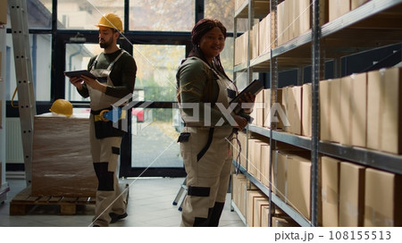 Portrait of joyful warehouse manager holding digital tablet, checking packaging, ensuring smooth operations while employee in background handles packaged goods in cardboard boxes ready to be shipped Portrait of joyful warehouse manager holding digital tablet, checking packaging, ensuring smooth operations while employee in background handles packaged goods in cardboard boxes ready to be shipped 108155513