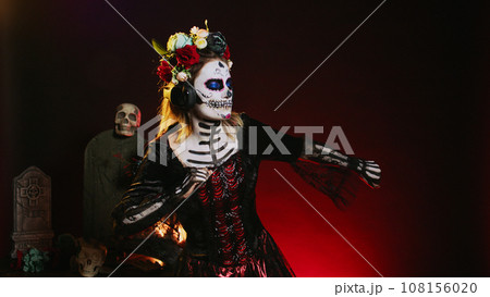 La cavalera catrina listening to music on audio headset, using headphones to enjoy mp3 song over black background. Creepy horror woman using headphones on holy dios de los muertos tradition. La cavalera catrina listening to music on audio headset, using headphones to enjoy mp3 song over black background. Creepy horror woman using headphones on holy dios de los muertos tradition. 108156020