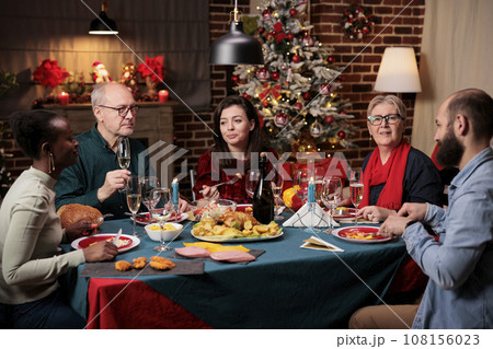 Old person at festive dinner with friends enjoying christmas celebration at home, place filled with decorations and xmas tree. Grandma preparing traditional food for holiday season event. 108156023