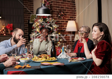 Family raising wine glasses for toast during festive dinner, enjoying holiday time at home with traditional meal. Diverse people giving speech to thank everyone for gathering around the table. 108156078