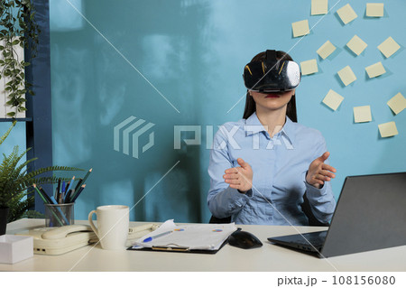 Female corporate employee with virtual reality glasses to visualize work project. Professional woman with immersive multimedia viewer working at office desk gesturing with hands. Female corporate employee with virtual reality glasses to visualize work project. Professional woman with immersive multimedia viewer working at office desk gesturing with hands. 108156080