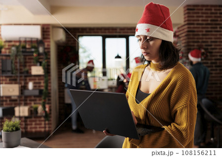 Woman project manager wearing santa hat managing tasks on laptop while standing in winter festive office. Employee holding portable computer and working for deadline at christmas eve 108156211