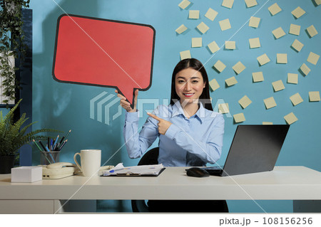 Professional asian woman with advertising speech bubble board against blue wall, showing isolated cardboard text message. Corporate employee holding poster with copy space. 108156256