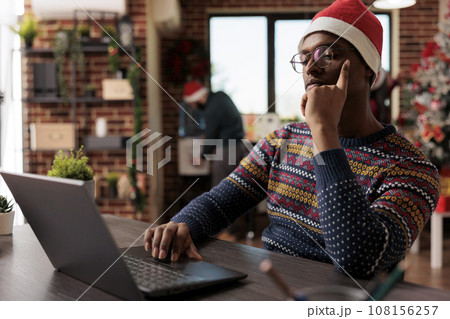 Startup company african american man employee in santa hat working on laptop in office at christmas holiday. Worker thinking while managing project report at festive decorated workplace 108156257