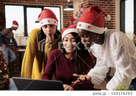 Young women colleagues wearing santa hats discussing and collaborating on tasks on christmas. Diverse smiling coworkers team working on laptop together in decorated office on xmas eve Young women colleagues wearing santa hats discussing and collaborating on tasks on christmas. Diverse smiling coworkers team working on laptop together in decorated office on xmas eve 108156265