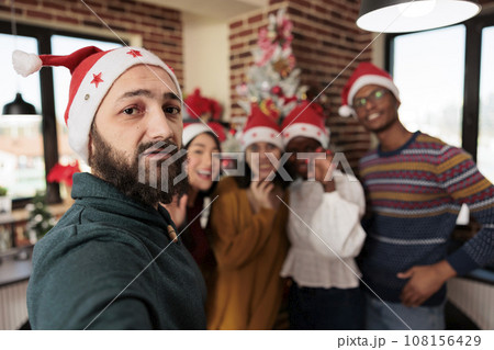 Diverse workers taking smartphone selfie at christmas corporate party in decorated office. Colleagues in santa hats posing for mobile phone photo while celebrating new year at workplace 108156429