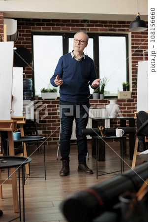 Senior man wearing glasses teaching art to adults in group class, standing in classroom introducing to drawing during creative workshop. Learning to draw from proffesional instructor Senior man wearing glasses teaching art to adults in group class, standing in classroom introducing to drawing during creative workshop. Learning to draw from proffesional instructor 108156660