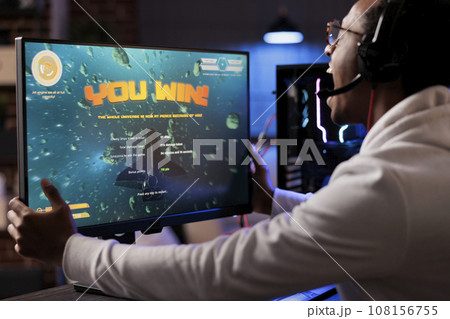 Ecstatic gamer playing intense classic arcade spaceship racing videogame, shaking computer display and yelling after winning. African american man having burst of happiness after game victory Ecstatic gamer playing intense classic arcade spaceship racing videogame, shaking computer display and yelling after winning. African american man having burst of happiness after game victory 108156755