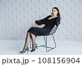 Fashion portrait of a stylish brunette girl wearing a black dress. Woman with long hair wearing red earrings. Model posing standing at the chair. Posing in white studio 108156904