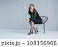 Fashion portrait of a stylish brunette girl wearing a black dress. Woman with long hair wearing red earrings. Model posing standing at the chair. Posing in white studio 108156906