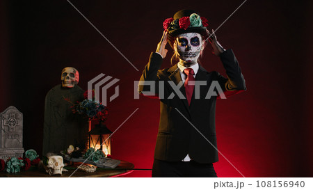 Cavalera catrina with suit and hat looking mysterious wearing festival skull body art, santa muerte celebrating dios de los muertos in studio. Woman looking like holy mexican entity. Cavalera catrina with suit and hat looking mysterious wearing festival skull body art, santa muerte celebrating dios de los muertos in studio. Woman looking like holy mexican entity. 108156940