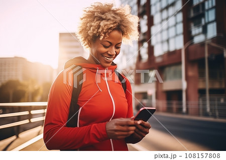 Sporty mature African American woman in sports outfit with smartphone while training outdoor. Slender black lady resting after jogging or walking in city street. Active lifestyle in urban environment. 108157808
