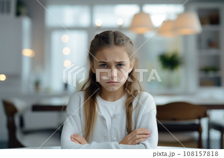 Portrait of a little sullen girl in a home setting, folding her arms and frowning eyebrows. Cute little girl is upset and offended. Beautiful child expresses negative emotion with face and pose. 108157818