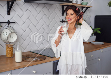 Charming beautiful woman drinks fresh milk at the morning in stylish kitchen. Beautiful girl is preparing breakfast at modern cozy kitchen. 108162490