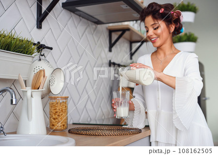 Beautiful smiling woman pours fresh milk from a carafe into a glass, in the stylish cozy kitchen at the morning. Healthy eating and diet concept. 108162505