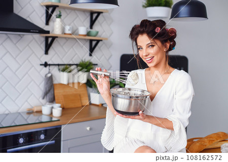 Attractive girl in white bathrobe kneads a dough in a metal plate at stylish modern kitchen. Young pretty woman baking on home kitchen. 108162572