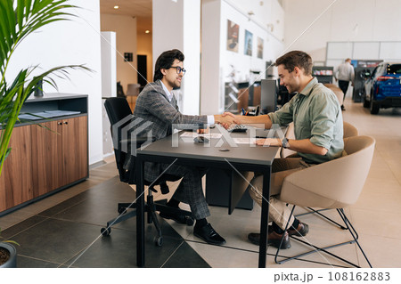 Wide shot of happy client male purchasing automobile in dealership, signing paper, shaking hands with car dealer in suit enjoying successful agreement. Concept of buying new auto at showroom. 108162883